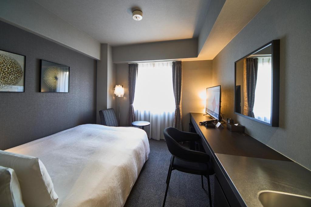A modern hotel room with a large bed, a desk and chair, a wall-mounted TV, and a window with curtains letting in natural light. The room has neutral decor with framed artwork on the walls.