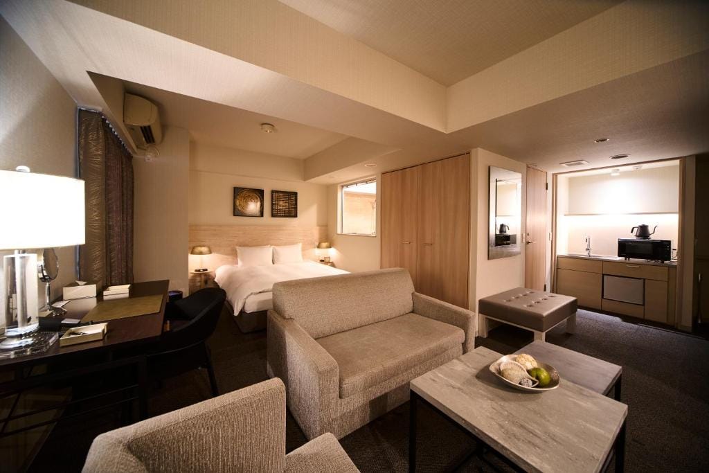 A modern hotel suite with a double bed, a sofa and coffee table, a desk with a lamp, and a kitchenette featuring a microwave, kettle, and sink. Neutral colors and soft lighting create a cozy atmosphere.
