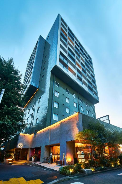 A modern, multi-story hotel building with an overhanging upper section, large windows, exterior lighting, and a restaurant at street level, surrounded by trees at dusk.