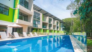 Modern apartment building with green accents, large windows, and balconies overlooks a long outdoor swimming pool lined with lounge chairs and umbrellas, surrounded by trees and greenery under a clear sky.