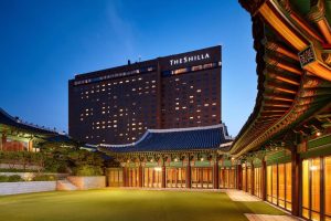 A modern high-rise hotel labeled “The Shilla” stands behind a traditional Korean palace-style building with ornate, colorful roofs and large windows, illuminated at dusk.