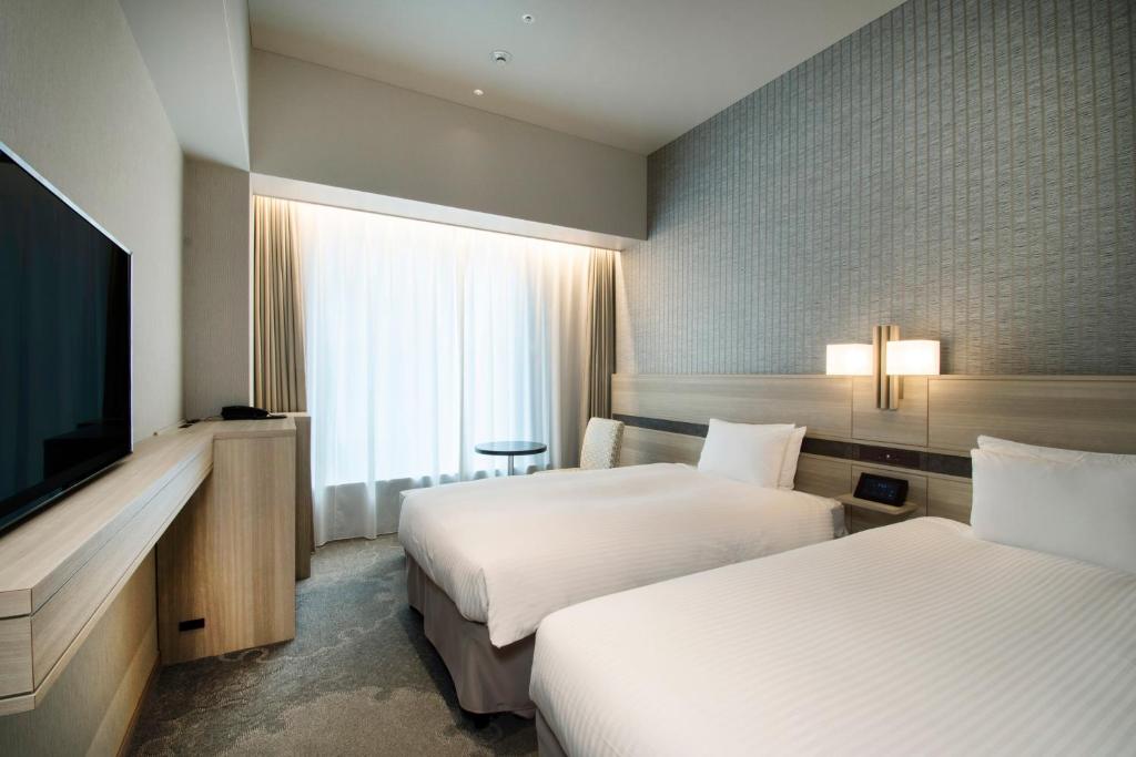 Modern hotel room with two neatly made single beds, a large flat-screen TV on a wooden console, a small round table with a chair by a large window covered with sheer curtains, and soft ambient lighting.