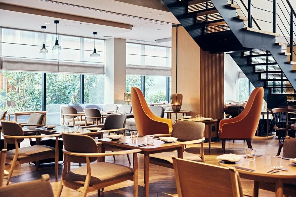 Modern restaurant interior with wooden tables and chairs, two tall orange armchairs, large windows letting in natural light, and a black metal staircase leading upstairs. The atmosphere is bright and airy.