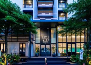 Modern hotel entrance with large glass doors and a sign reading Cross Vibe Bangkok Sukhumvit. Surrounded by green trees and plants, the building features balconies and a well-lit lobby area inside.