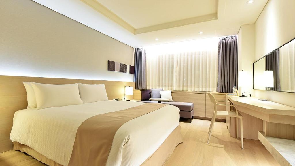 A modern hotel room with a large bed, beige bedding, a desk with a chair, a sofa by the window, and soft lighting creating a warm, inviting atmosphere.