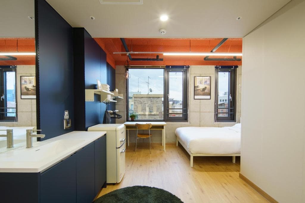 Modern, minimalist studio apartment with a bed by large windows, a desk and chair, a small fridge, and a white sink area in the foreground. Wooden flooring and industrial-style decor complete the space.