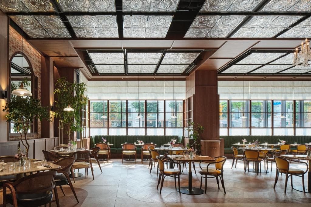 Elegant restaurant interior with vintage-style decor, patterned ceiling panels, large windows, greenery, and wooden tables and chairs arranged neatly in a spacious, well-lit dining area.