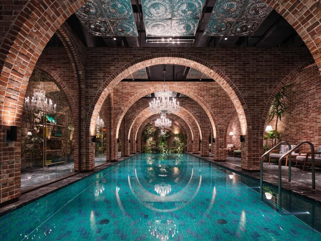 An indoor pool with turquoise tiles, surrounded by brick arches and walls, crystal chandeliers reflecting on the water, and lush green plants creating a luxurious, elegant atmosphere.