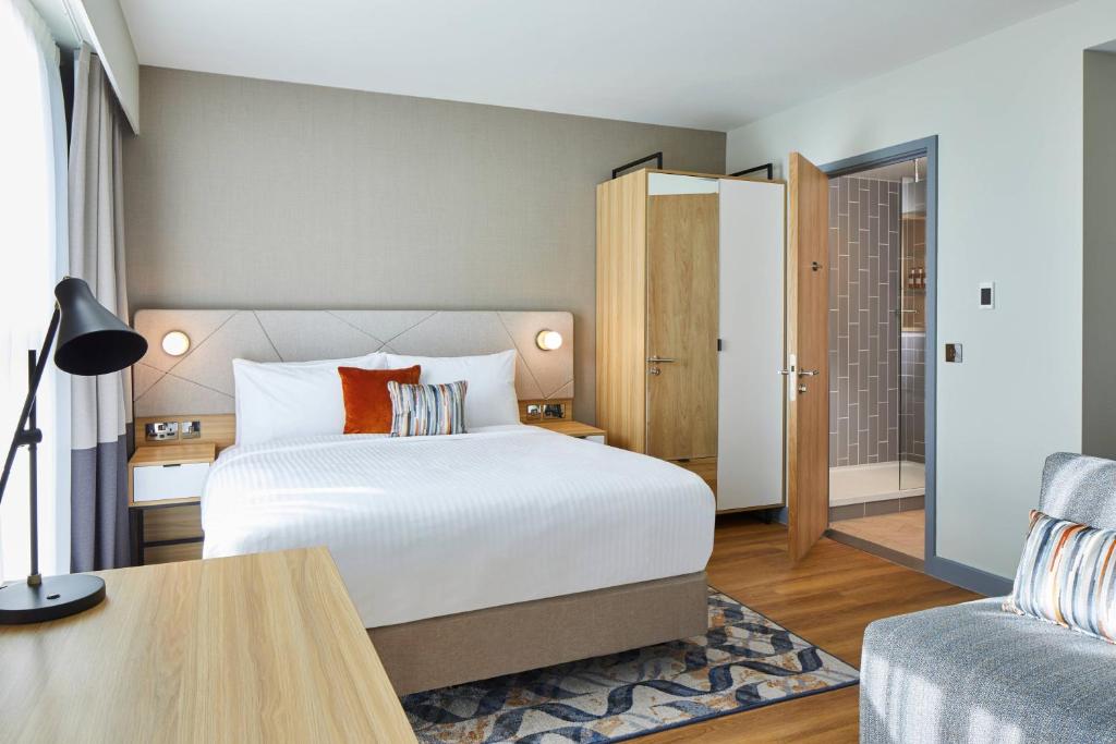 A modern hotel room with a neatly made double bed, wooden furniture, a desk lamp, a wardrobe with an open door, and a view into a tiled bathroom. A patterned rug and decorative pillows add color.