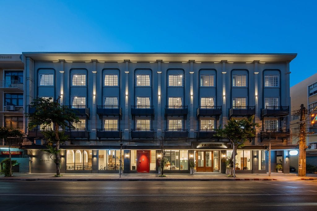 A modern three-story building with large windows evenly lit at dusk, featuring a bright red door at the center and shops or restaurants at street level; trees line the sidewalk in front.