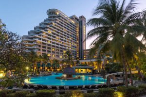 A large, modern hotel with tiered balconies overlooks a tropical outdoor swimming pool, surrounded by palm trees, sun loungers, and evening lighting at sunset.