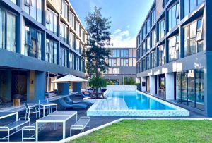 Modern apartment complex with two buildings facing each other, featuring a central outdoor swimming pool, lounge chairs, umbrellas, tables, and a grassy area under a bright blue sky.