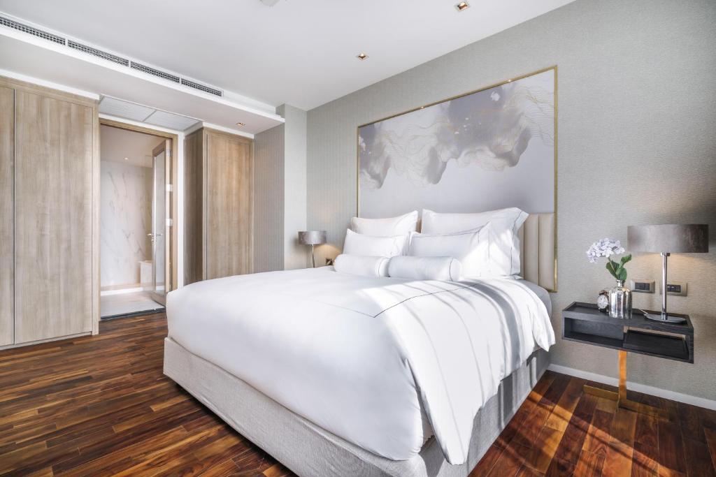 A modern, bright hotel room with a large neatly made bed, white bedding, wooden floor, bedside tables with lamps, a flower vase, and a large abstract artwork above the bed. Wardrobe and hallway are visible in the background.