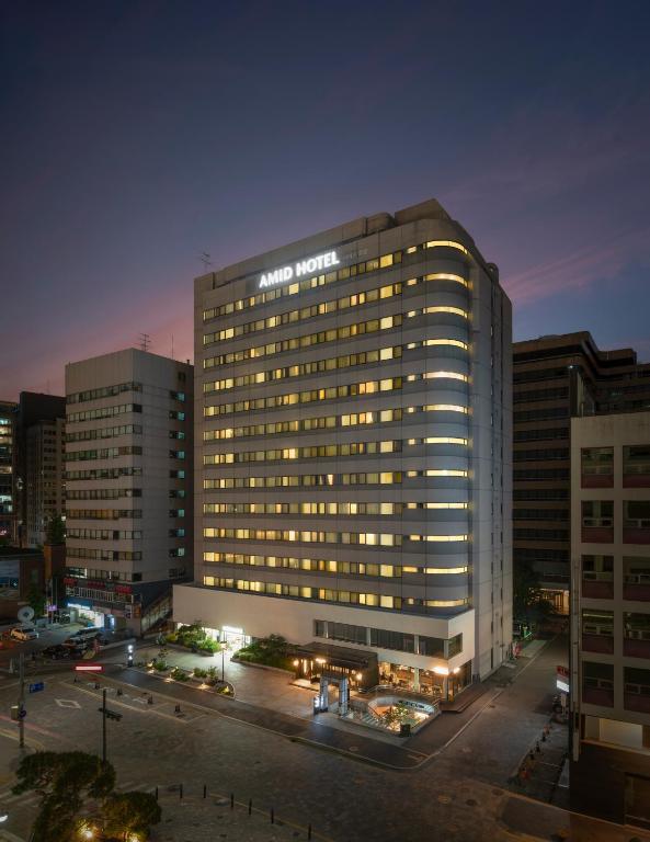 A tall, modern hotel building labeled AMID HOTEL is illuminated at dusk, with lights on in many windows. The hotel sits on a street corner in an urban area, surrounded by other buildings.
