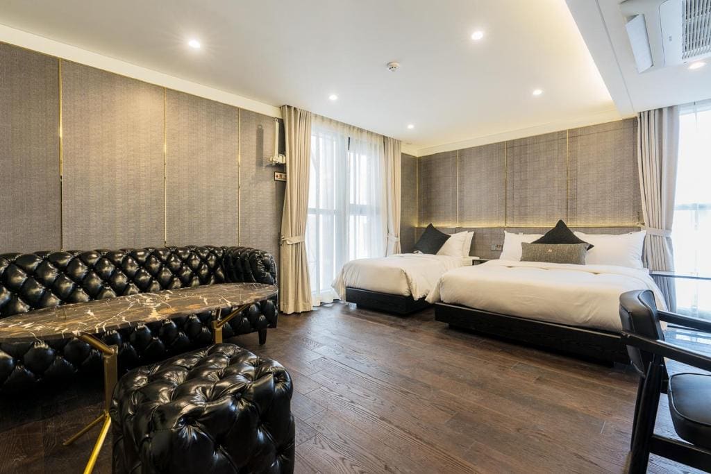 A modern hotel room with two large beds, a black tufted leather sofa, a marble coffee table, a matching ottoman, wooden floors, large windows with light curtains, and soft overhead lighting.