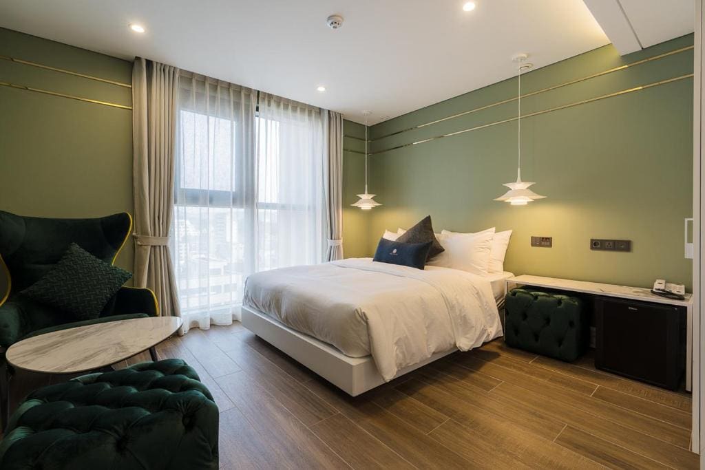 A modern hotel room with a double bed, green walls, wood flooring, velvet green chairs, a small round marble table, two pendant lights, large window with sheer curtains, and a mini fridge beside the bed.