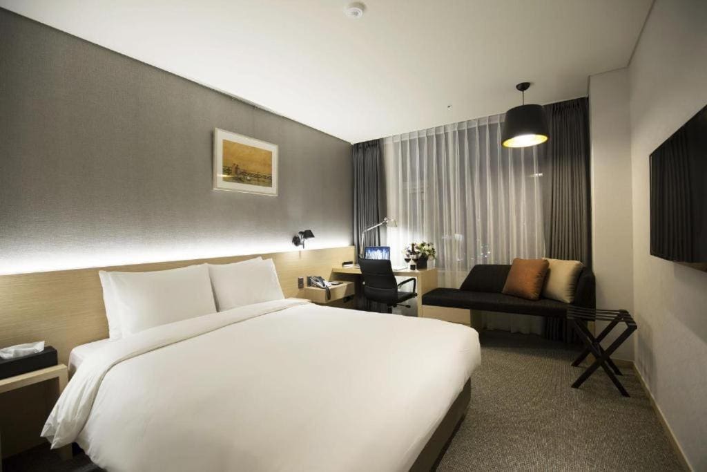 A modern hotel room with a large bed, white bedding, a desk with a chair and lamp, a small sofa with cushions, a wall-mounted TV, and a window with sheer curtains.