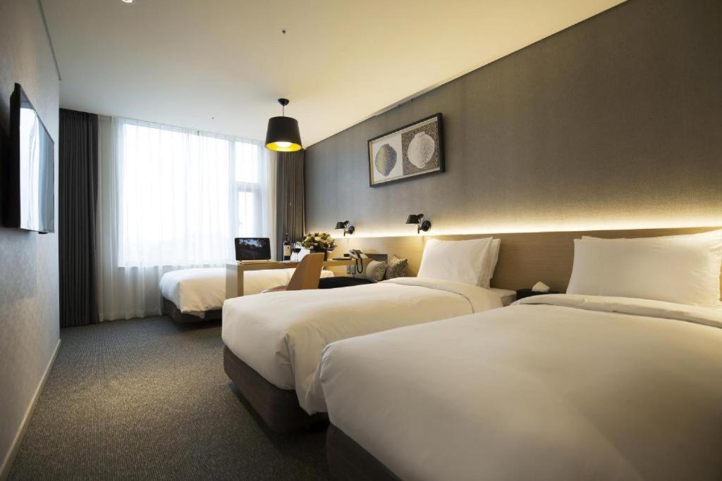 A modern hotel room with three neatly made single beds, a desk with a chair and lamp by a large window, and contemporary wall art above the beds. The room has neutral colors and soft lighting.
