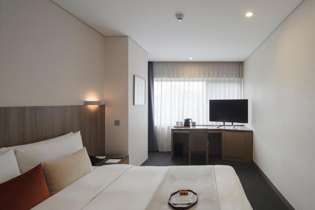A modern hotel room with a neatly made bed, two accent pillows, a bedside table, a desk with a chair, a flat-screen TV, a kettle, and a plate with snacks near a large window with sheer curtains.