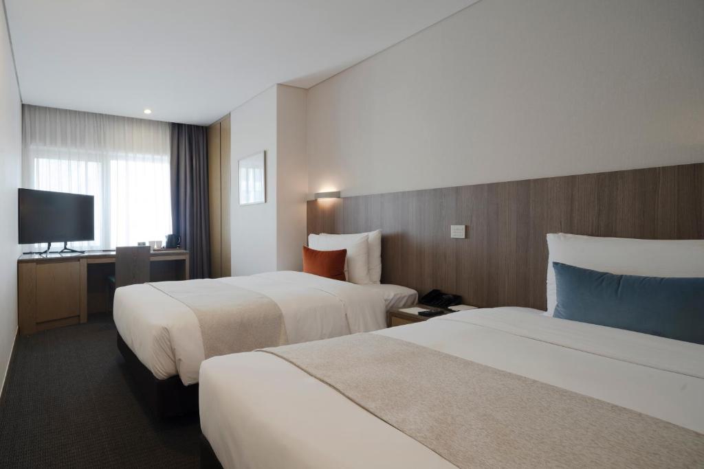 A modern hotel room with two neatly made single beds, each with accent pillows, a desk with a chair, a flat-screen TV, and a large window with sheer curtains letting in natural light.