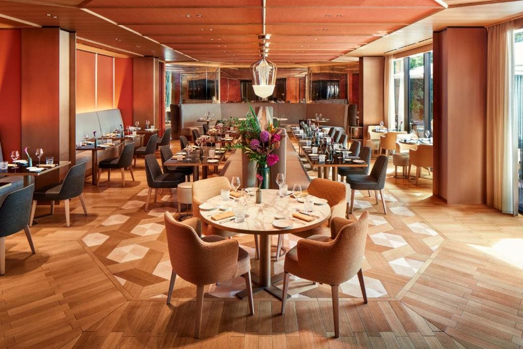 Elegant restaurant interior with wooden floors, orange and gray chairs, marble tables set with glassware, and a floral centerpiece. Warm lighting and large windows create a welcoming, upscale atmosphere.