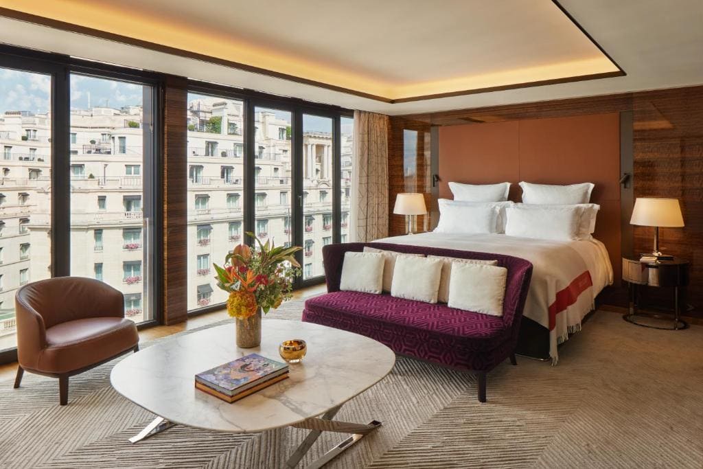 A modern hotel room with floor-to-ceiling windows, a large bed, a purple sofa at the foot of the bed, a round marble coffee table with flowers and books, and a brown armchair. City buildings are visible outside.