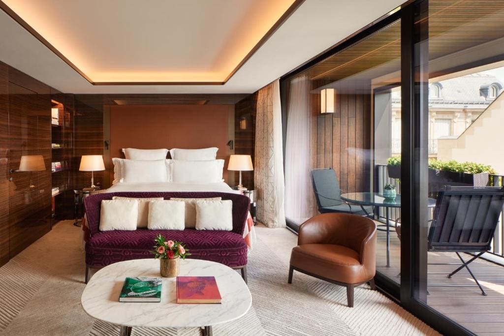 A modern hotel room with a large bed, two bedside lamps, a purple sofa, a round marble coffee table with books and flowers, a brown armchair, and a balcony with outdoor seating and potted plants.