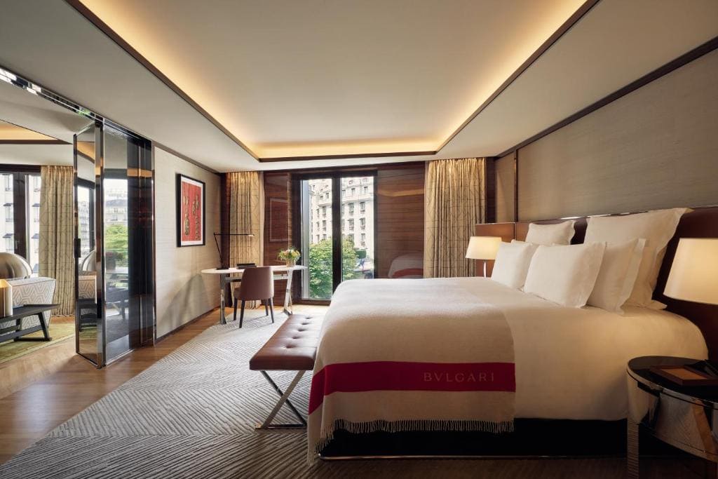 Modern hotel room with a large bed, leather bench, desk near a window, and neutral decor. Floor-to-ceiling windows offer a city view. Soft lighting and elegant furnishings create a luxurious, inviting atmosphere.