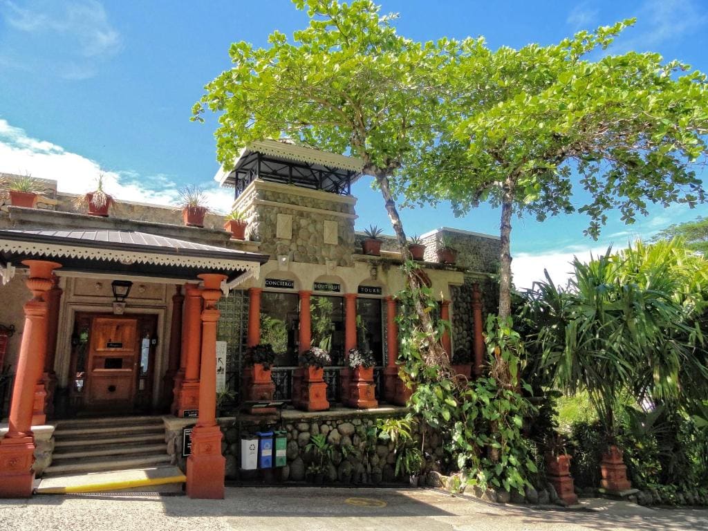 A stone and brick building with red columns and lush green plants, including a large tree in front, under a bright blue sky with scattered clouds. Steps lead to the entrance and potted plants line the porch.