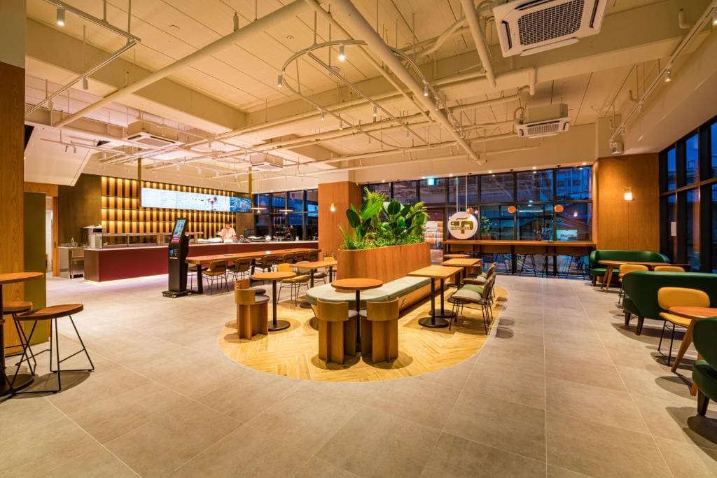 Spacious, modern cafe interior with wooden tables, green chairs, a central planter with large plants, and floor-to-ceiling windows showing a cityscape at dusk. Warm lighting and a welcoming atmosphere.