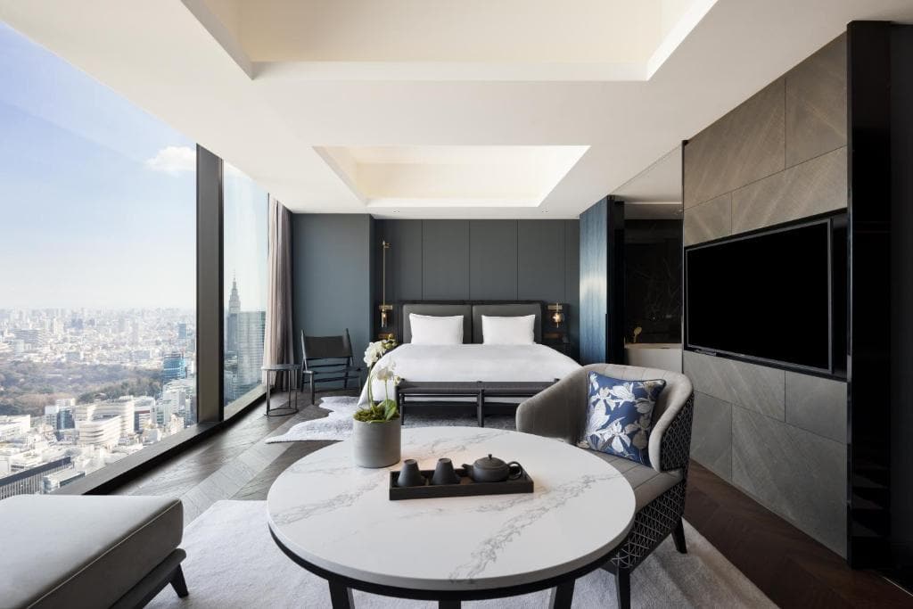 A modern hotel room with a large bed, floor-to-ceiling windows offering a city view, a marble coffee table with a tea set, an armchair, and a wall-mounted flat-screen TV.