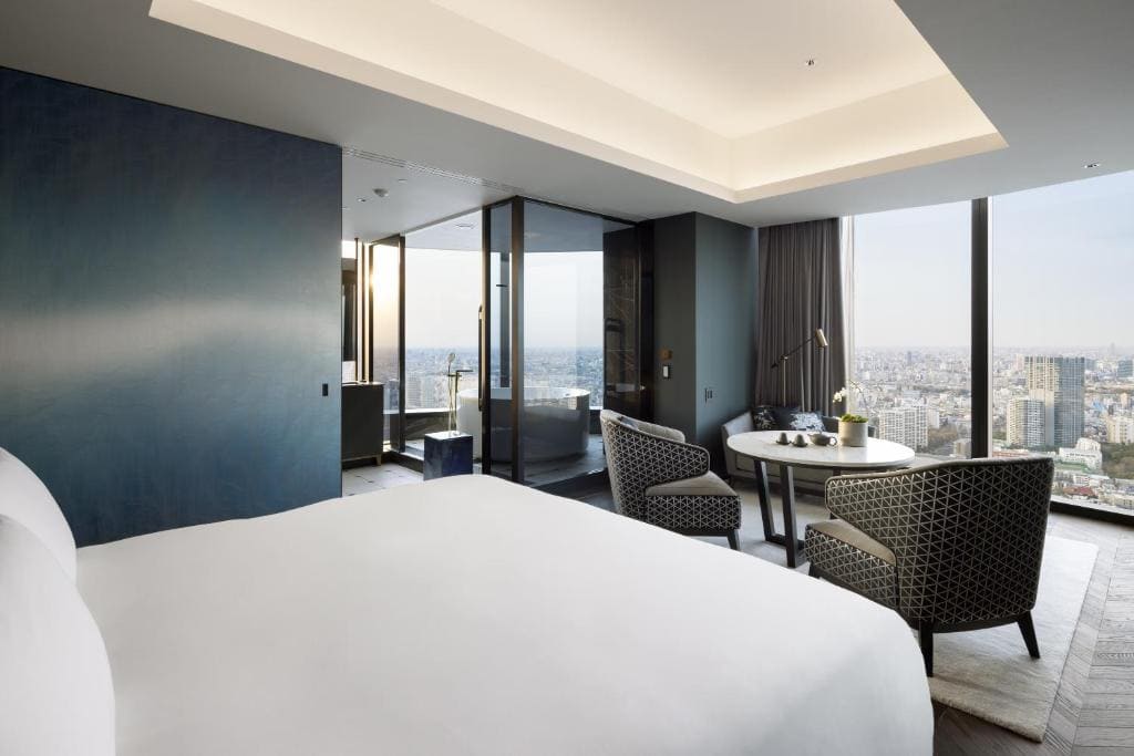 A modern hotel room with a large bed in the foreground, two patterned chairs and a round table by floor-to-ceiling windows, overlooking a cityscape with tall buildings and a distant skyline.