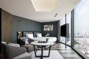 A modern hotel room with large floor-to-ceiling windows overlooking a cityscape, featuring a bed, a seating area with two armchairs, a small round table, a wall-mounted TV, and minimalist decor.