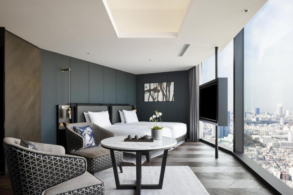 A modern hotel room with large floor-to-ceiling windows overlooking a cityscape, featuring a bed, a seating area with two armchairs, a small round table, a wall-mounted TV, and minimalist decor.