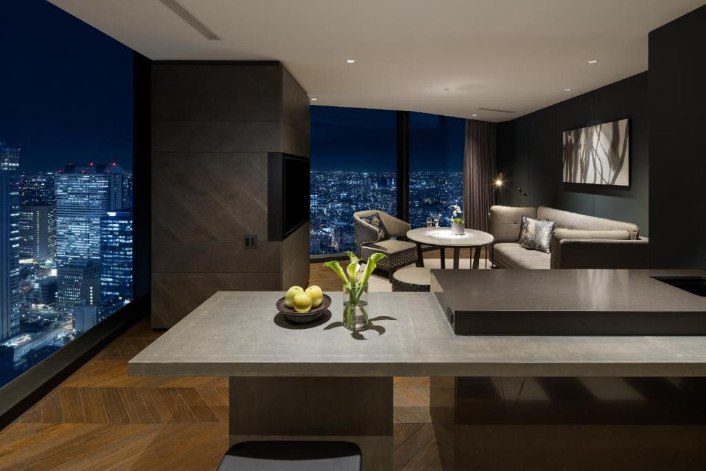 Modern hotel suite at night with city view, featuring a sleek kitchen island, a vase with flowers, a bowl of green apples, a sofa, round table, armchairs, and floor-to-ceiling windows overlooking illuminated skyscrapers.