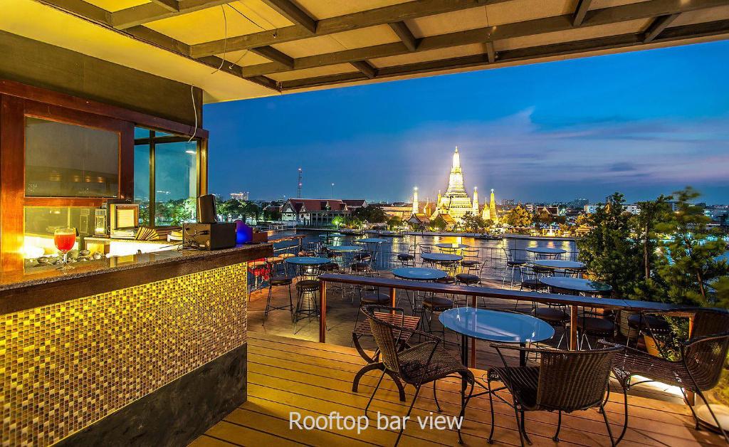 Rooftop bar with tables and chairs overlooking a river at sunset, featuring a beautifully lit temple and city skyline in the background. Rooftop bar view appears at the bottom of the image.