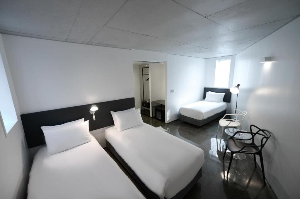 A minimalist, modern hotel room with two single beds side by side, a third single bed against the wall, white walls, gray floors, a small desk with two chairs, and simple lighting.