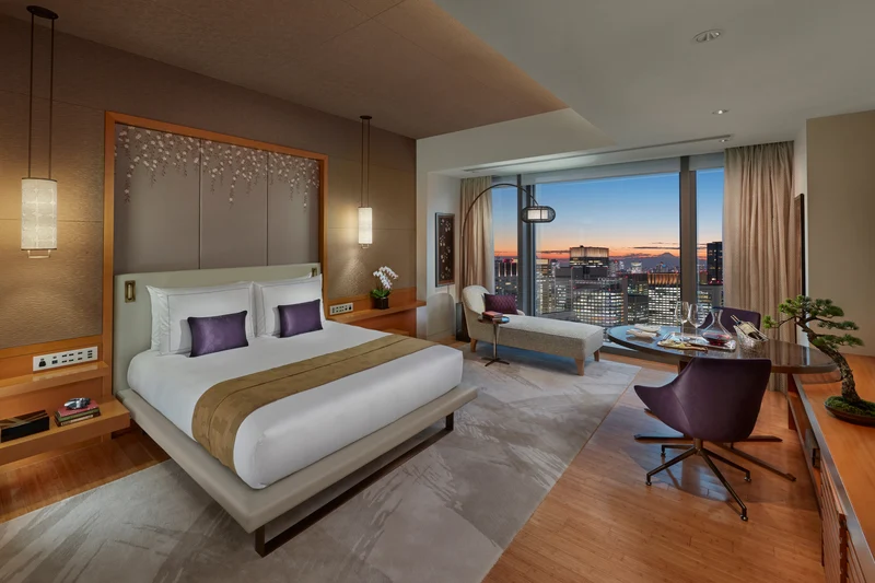 Luxurious hotel room with a large bed, modern furniture, a small dining area, and a floor-to-ceiling window offering a city view at sunset. The room features warm lighting and elegant decor.