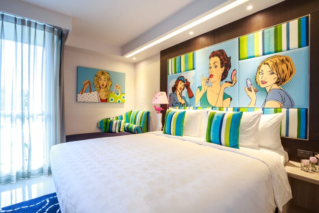 A modern hotel room with a large bed, striped green and blue pillows, pop art paintings of women on the wall, a striped chair, a pink lamp, and a window with sheer curtains letting in natural light.