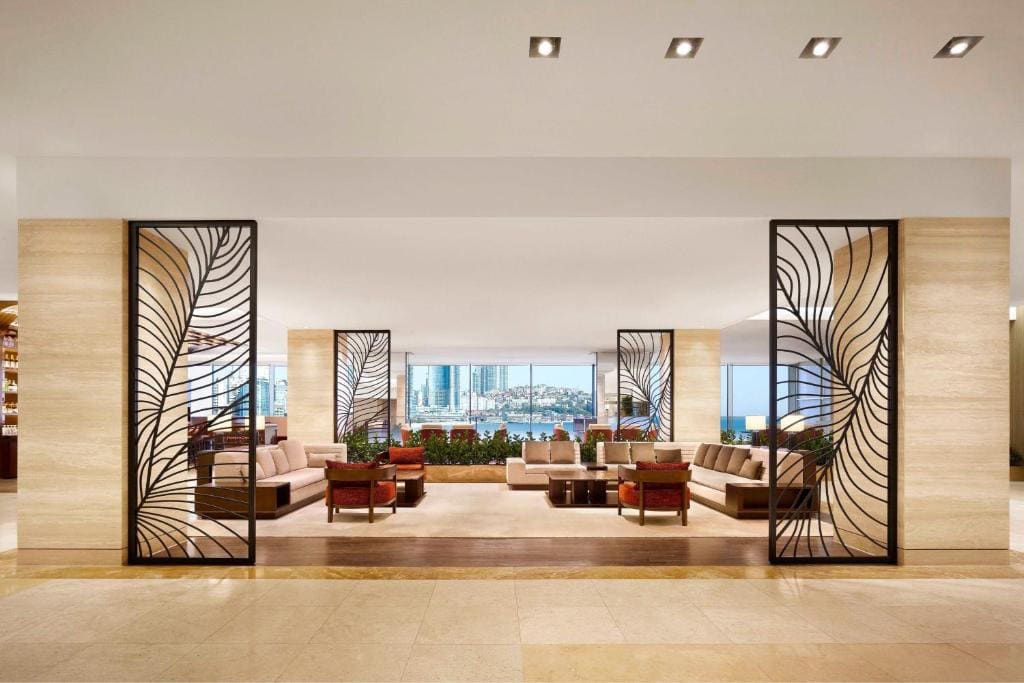 A modern hotel lobby with beige marble floors, elegant seating areas, decorative black metal screens, large windows, and a view of a city skyline in the background.