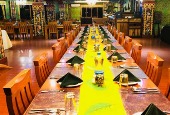 A long dining table set for a meal with wooden chairs, green napkins, glasses, and water bottles arranged neatly. The room is decorated with colorful patterns and has a warm, inviting atmosphere.