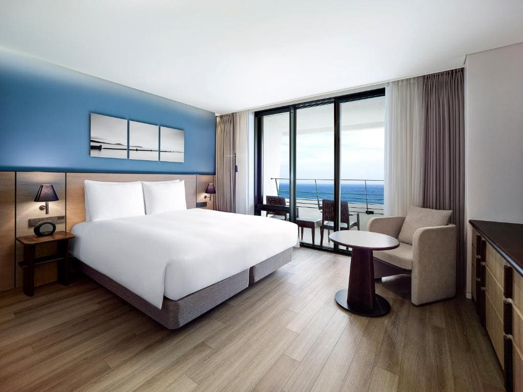 A modern hotel room with a large bed, two side tables, a blue accent wall with artwork, armchair, round table, and floor-to-ceiling windows opening to a balcony with an ocean view.