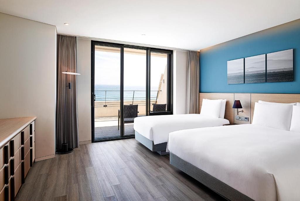Modern hotel room with two double beds, a blue accent wall, a desk, and a large sliding glass door opening to a balcony with an ocean view. Minimalist decor and natural light fill the room.