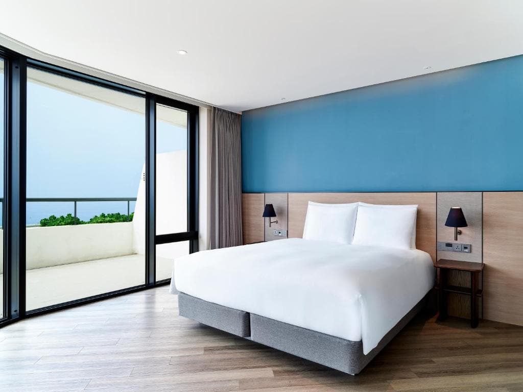 A modern hotel room with a large bed, blue accent wall, wooden floor, bedside tables with lamps, and floor-to-ceiling windows opening to a balcony with a view of the sea.
