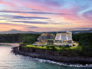 Modern seaside hotel complex perched on a rocky cliff, surrounded by lush greenery, overlooking the ocean at sunset with a colorful sky and distant mountains.