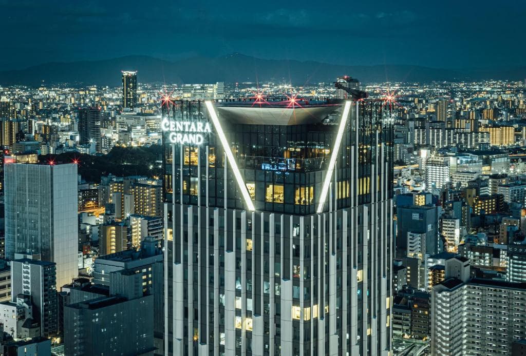 A tall, illuminated skyscraper with Centara Grand signage stands out against a densely packed cityscape at night, with many buildings and distant mountains visible under a dark blue sky.