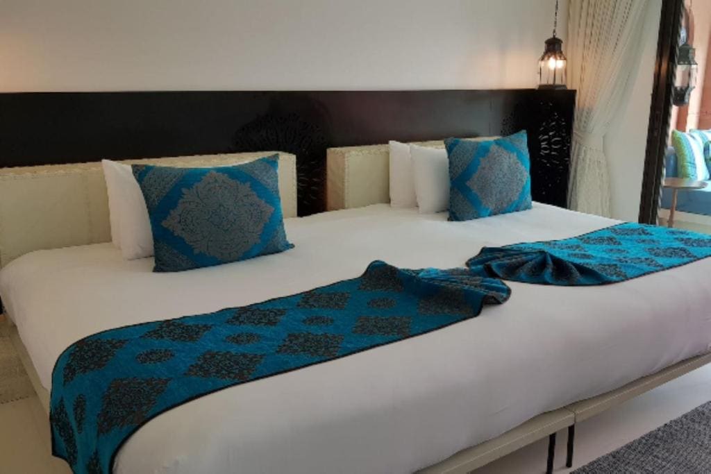 A neatly made bed with white sheets, blue decorative runners, and two matching blue patterned pillows. Black headboard, hanging lamp, and light streaming in from a window on the right.