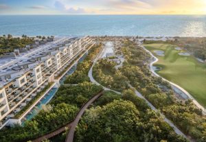 Aerial view of a modern beachfront resort with pools, lush gardens, a large golf course, and walkways along the coastline at sunset, overlooking the ocean.