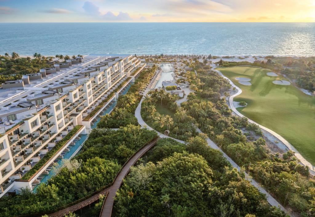 Aerial view of a modern beachfront resort with pools, lush gardens, a large golf course, and walkways along the coastline at sunset, overlooking the ocean.