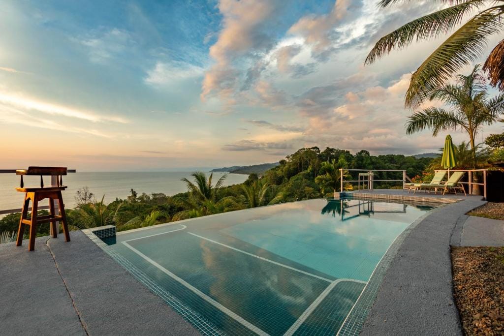 Infinity pool overlooking a lush tropical landscape and ocean at sunset, with palm trees, lounge chairs, and a wooden stool creating a relaxing atmosphere. The sky is partly cloudy with warm, golden light.